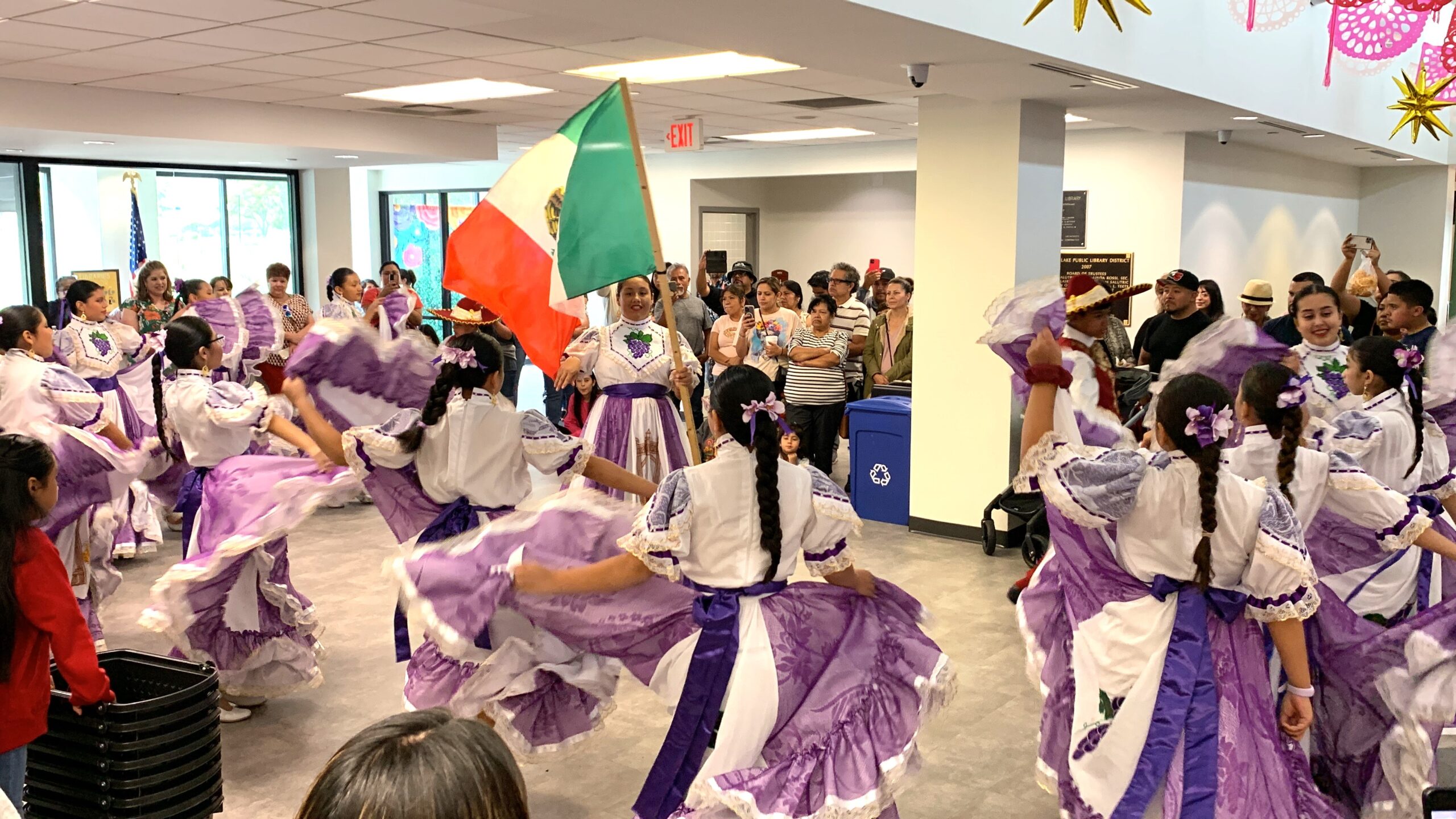 A group of folkloric dancers performing and waving the Mexican flag in the library lobby.